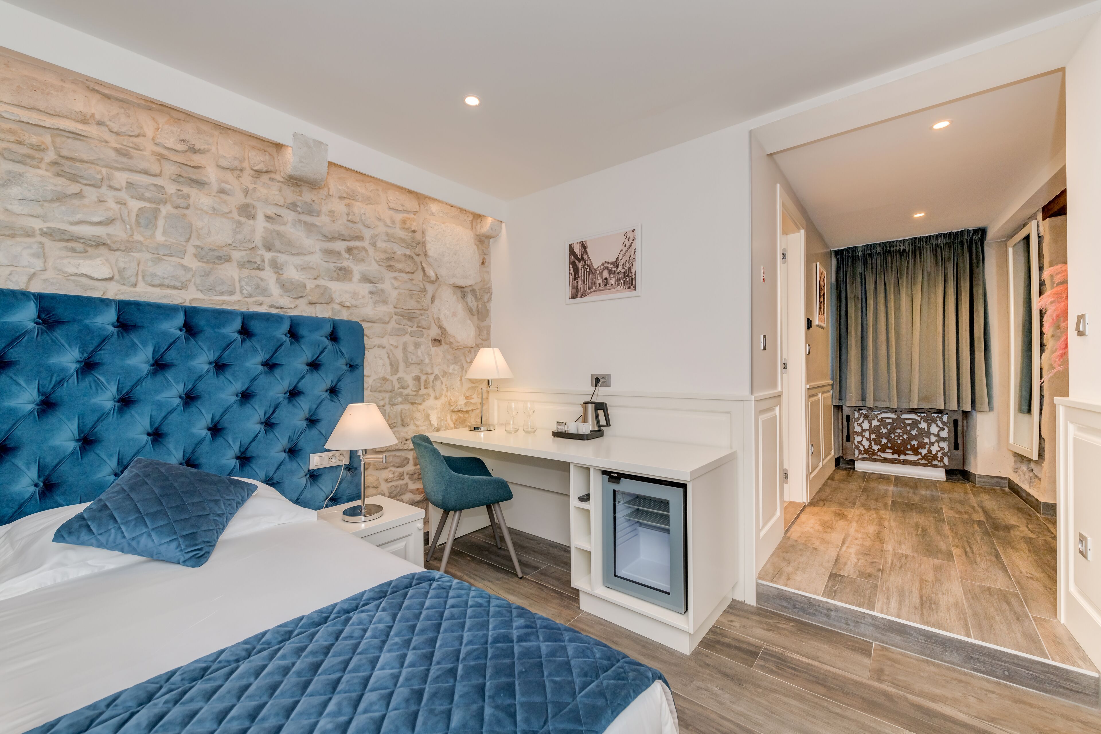 A bedroom with a stone wall, a tufted blue headboard, a desk with a chair, and a mini-fridge.