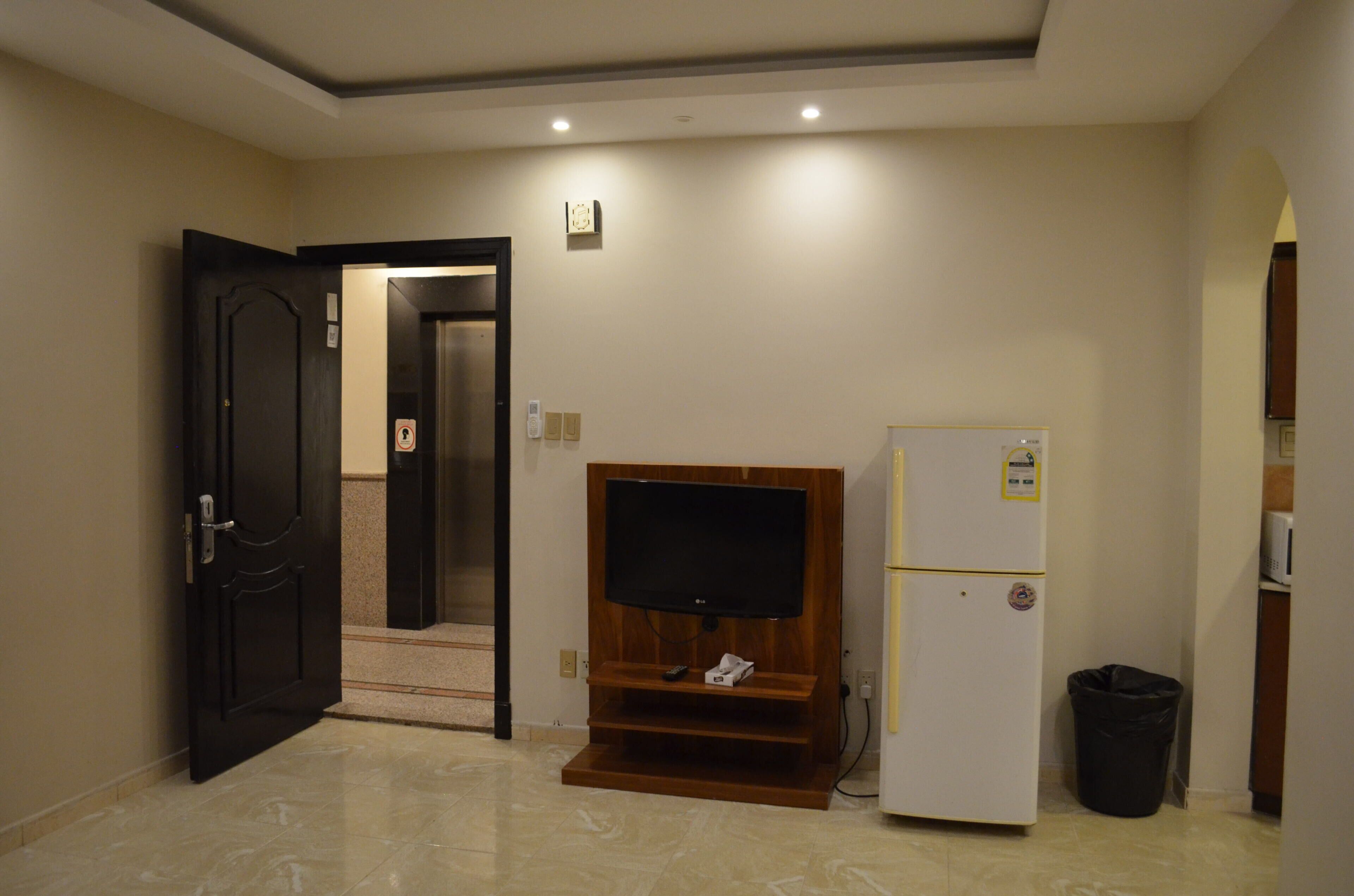 Habitación de hotel con una unidad de entretenimiento de madera, un refrigerador blanco, un basurero negro y una puerta que da a otra habitación.