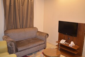Un salon avec un canapé marron, un fauteuil jaune, une table basse en verre, un téléviseur à écran plat fixé sur un meuble en bois, et un téléphone posé sur une étagère en bois.
