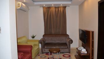 A living room with a brown sofa, a yellow armchair, a red armchair, a small round table, a TV on a wooden stand, and a telephone on a wooden shelf.