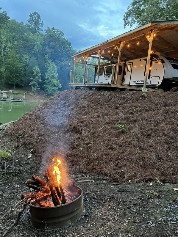 Un feu de camp avec des bûches et des braises, une structure en bois couverte et illuminée, et une étendue d’eau en arrière-plan.