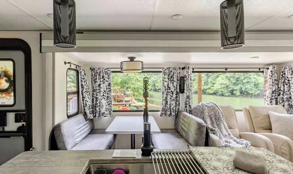 A cozy interior with a table and bench seating, large windows with curtains, and a view of greenery outside.