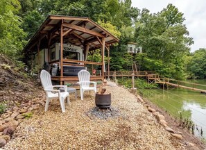 Een houten constructie met een overdekte veranda, een vuurkorf en twee witte stoelen.