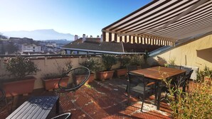 Una terraza en la azotea con mesa y sillas de madera, plantas en macetas y vistas a un paisaje urbano y montañas al fondo.