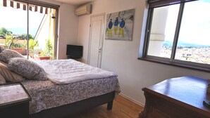 Un dormitorio con cama, escritorio, lámpara, televisor y ventana con vista a la ciudad.