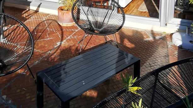 Un patio con una mesa y sillas, una planta en maceta y vista del interior a través de puertas de vidrio.