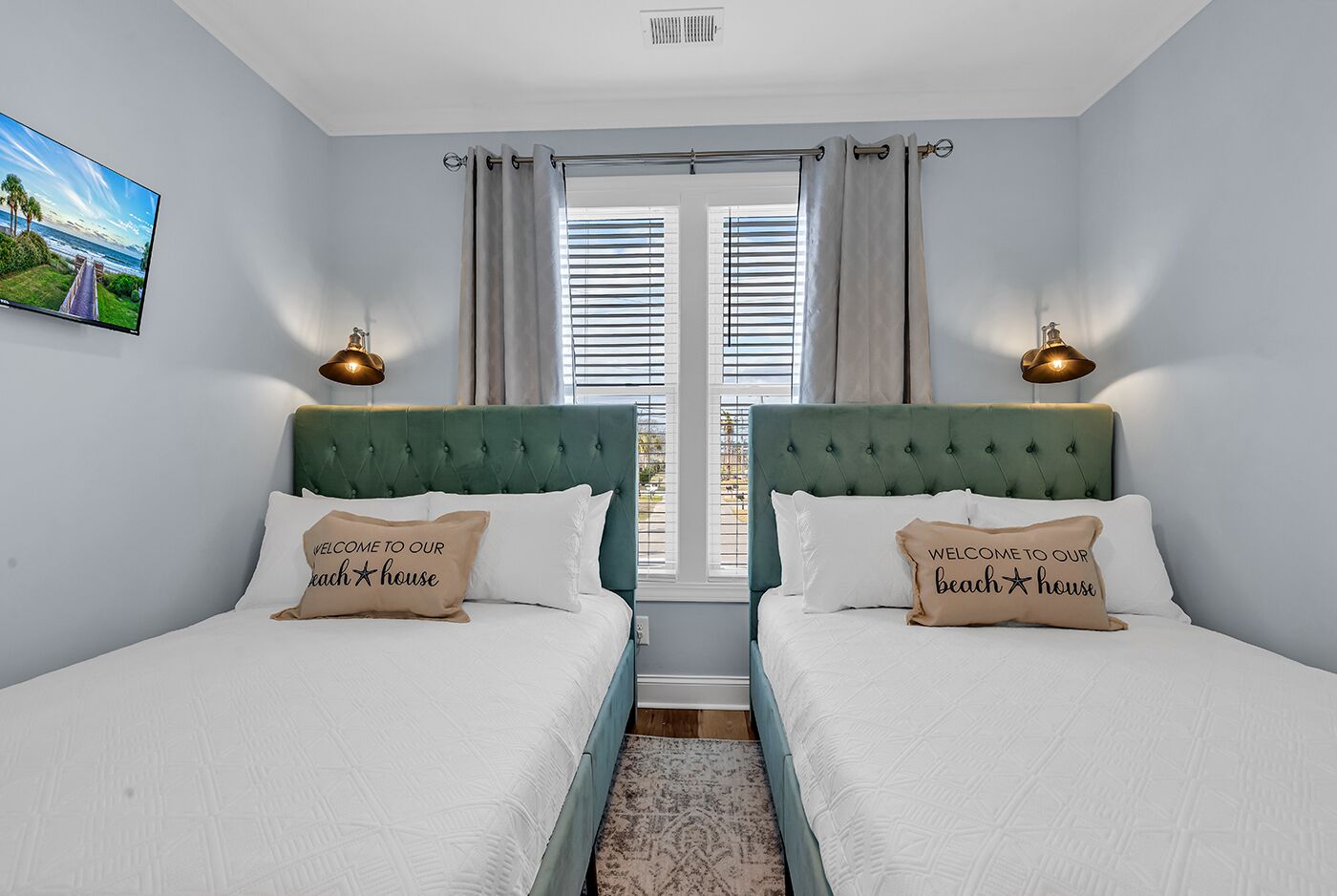 A bedroom with two beds, each with a green headboard, white bedding, and a pillow with "Welcome to our beach house" text. There are two wall-mounted lamps, a window with blinds, and a painting on the wall.