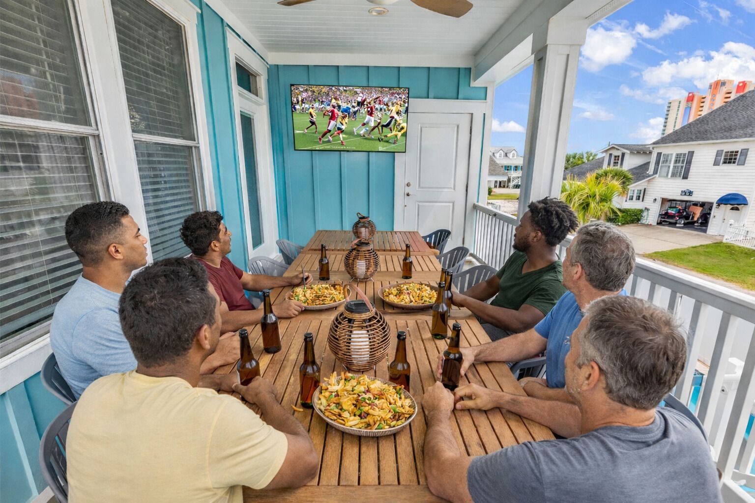 A group of people sitting at a wooden table on a porch with a TV displaying a sports game, surrounded by houses.