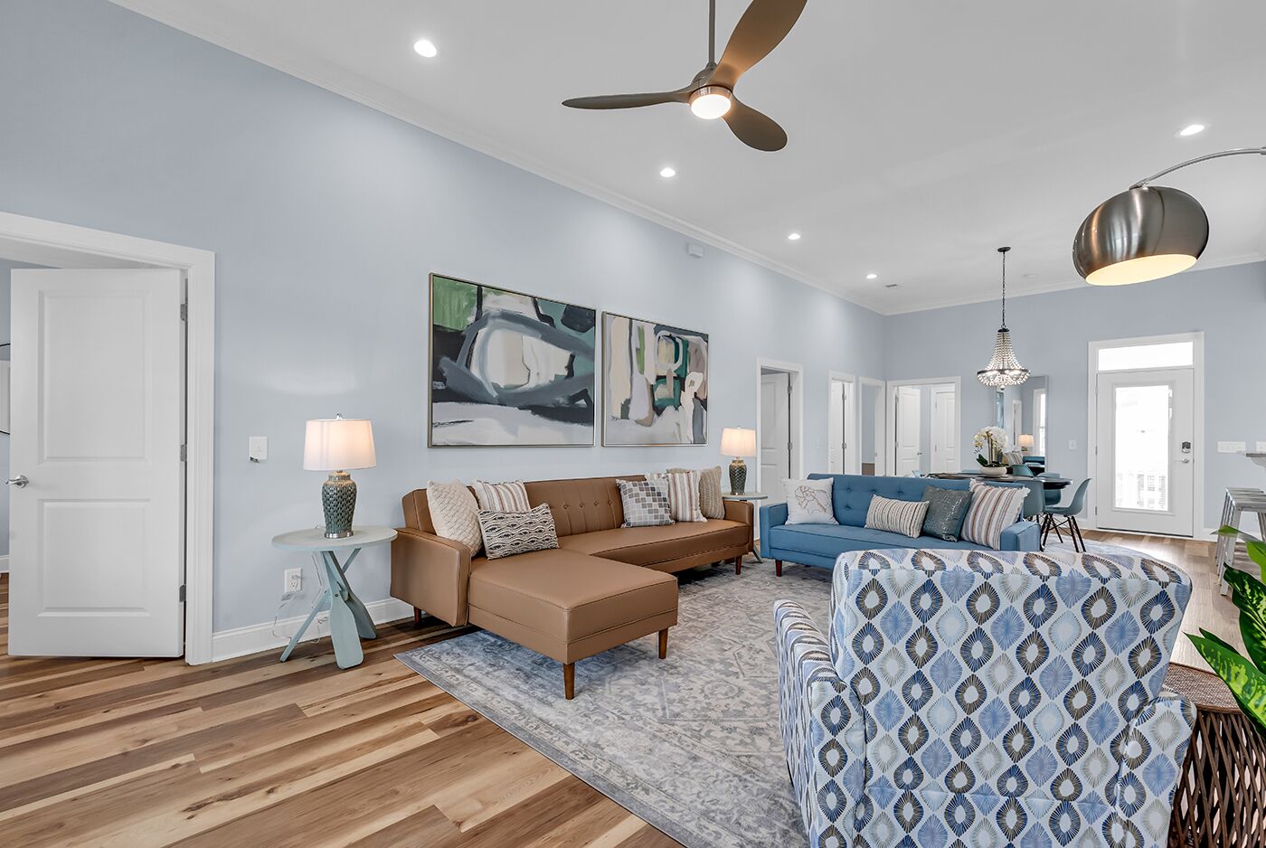 A spacious living room with a brown leather sofa, a blue patterned ottoman, and a blue sofa. There are two wall-mounted lamps, a ceiling fan, and a dining area visible in the background.