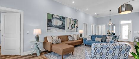 A spacious living room with a brown leather sofa, a blue patterned ottoman, and a blue sofa. There are two wall-mounted lamps, a ceiling fan, and a dining area visible in the background.