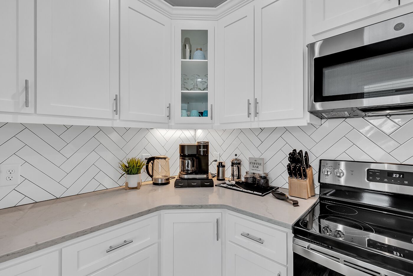 A modern kitchen with white cabinetry, a herringbone tile backsplash, a coffee station, and stainless steel appliances.