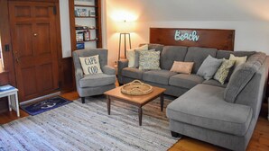 Un salon chaleureux avec un canapé gris, un petit fauteuil, une table basse en bois et une lampe. Un panneau indiquant « Plage » est accroché au mur, et une bibliothèque contient divers objets.