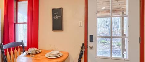 A dining area with a wooden table, two chairs, a glass of water, and a plate. A door with a window is visible in the background.