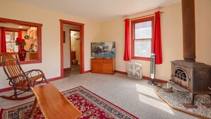 Un salon chaleureux avec un fauteuil à bascule, un téléviseur, une cheminée et une cuisine visible à travers une porte ouverte.
