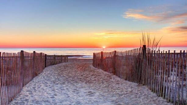 A sandy beach path leading to the ocean with a sunset in the background.