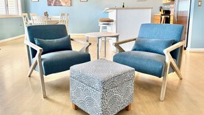 A modern living room with two blue armchairs, a patterned ottoman, a small round table, and a kitchen area in the background.
