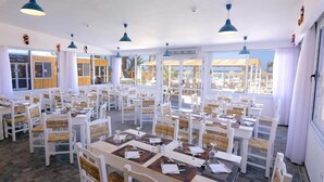 Zona pranzo con tavoli e sedie bianche, lampade a sospensione blu e ampie finestre vista spiaggia.