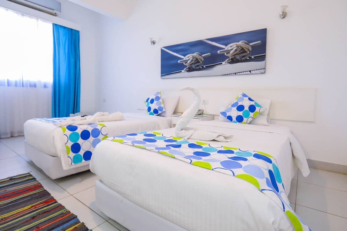 A hotel room with two beds, a large window with blue curtains, a painting of a ship on the wall, and a colorful striped rug.