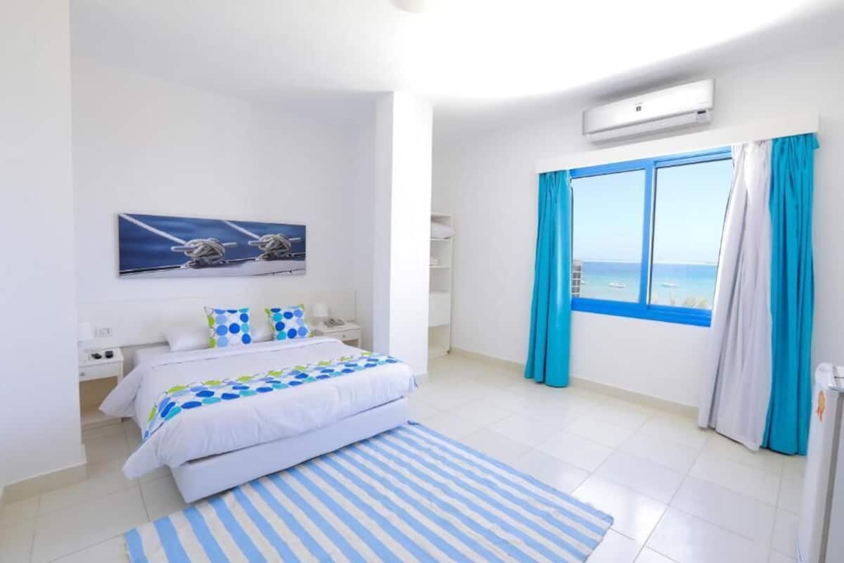 A modern bedroom with a large bed, a view of the sea through a window, and a painting of a sailboat.