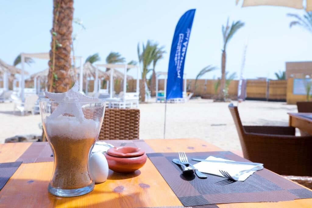 A table with a glass of iced coffee, a small bowl, and cutlery on a wooden table with a beach view in the background.