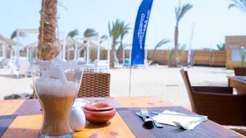 A table with a glass of iced coffee, a small bowl, and cutlery on a wooden table with a beach view in the background.