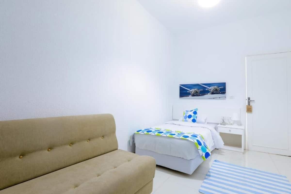A bedroom with a beige sofa, a bed with a blue and white polka dot bedspread, a white nightstand, and a wall-mounted picture.