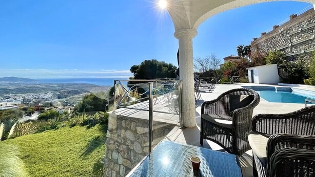 Une terrasse dotée d’une table en verre et de chaises en osier, offrant une vue sur une piscine et un paysage pittoresque de la campagne environnante.
