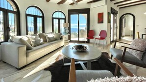Un salon spacieux avec un grand canapé, une table basse ronde et un ventilateur de plafond. De grandes fenêtres à arc permettent à la lumière naturelle d’inonder la pièce.