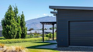 A modern building with a garage, a covered patio, and a well-maintained lawn with a mountain backdrop. - Hoheria Mountain Lodge (Wānaka)