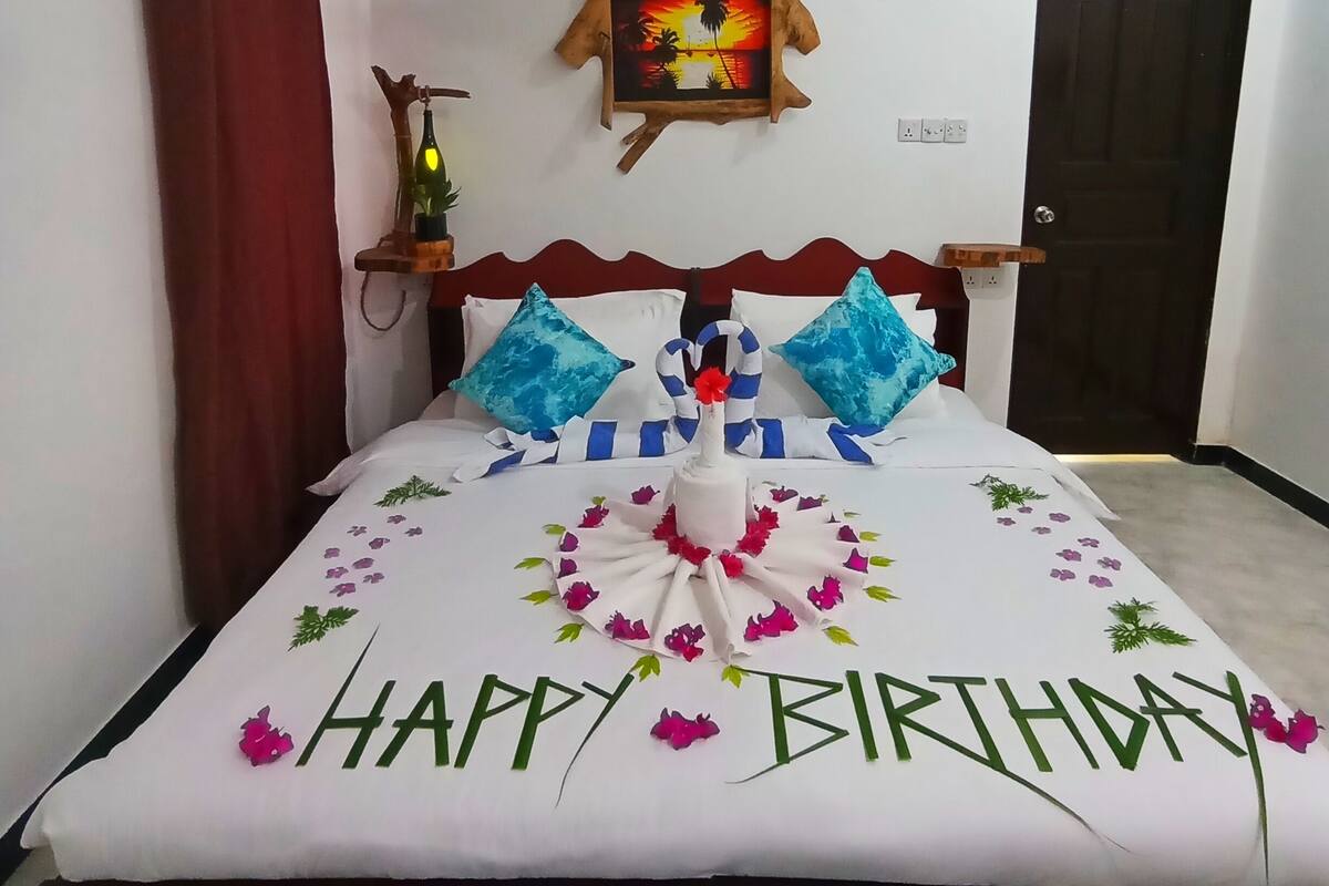 Una cama bien hecha con un mensaje de 'Feliz Cumpleaños', un adorno de pared y una puerta al fondo.