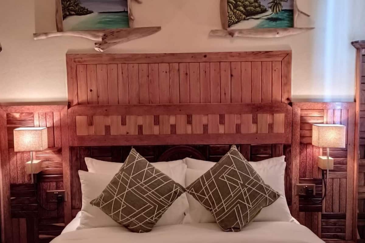 Una cama con estructura de madera, ropa de cama blanca y dos almohadas estampadas, flanqueada por mesitas de noche con lámparas, en una habitación con paredes de madera y dos cuadros colgados.