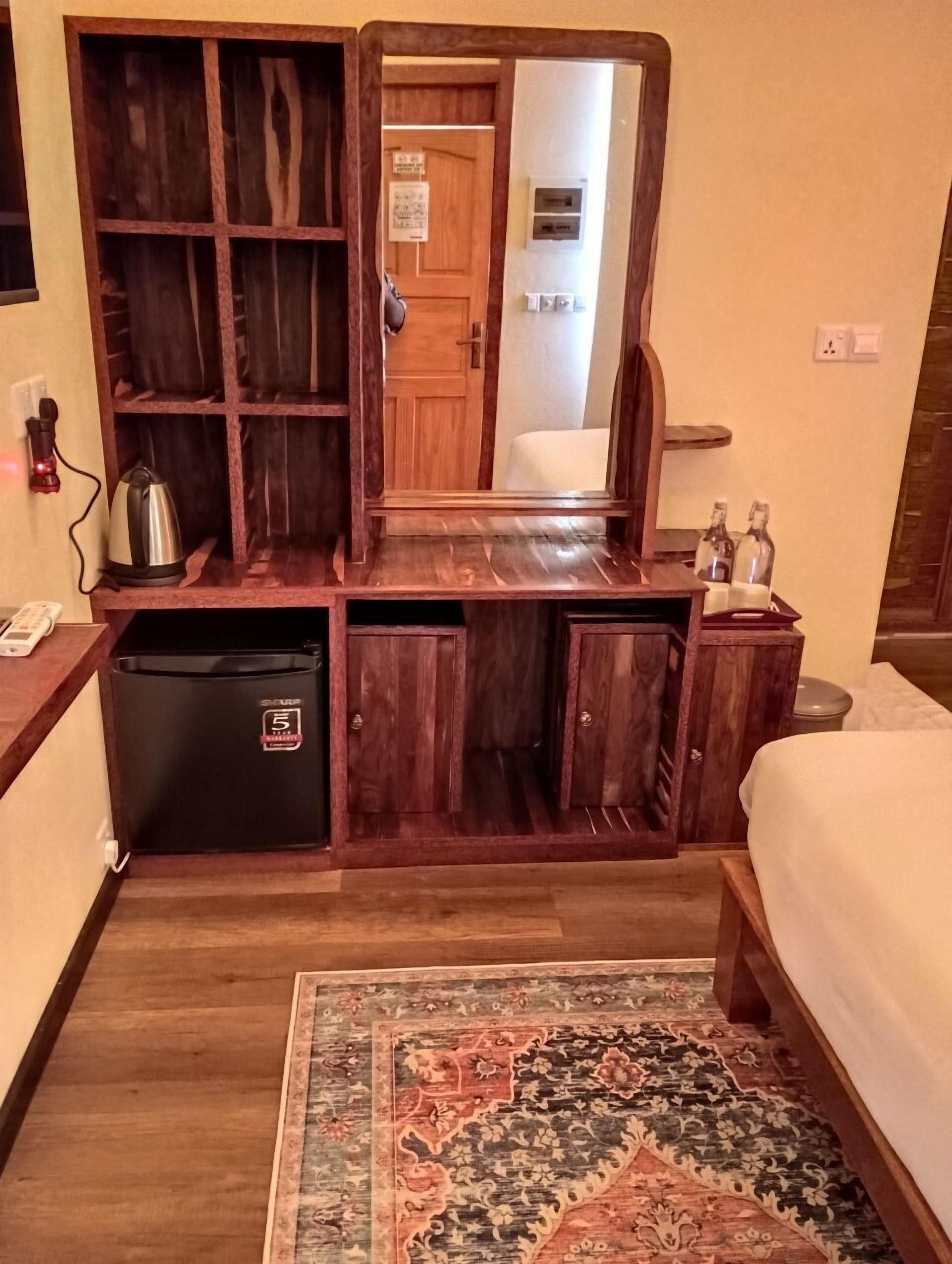A hotel room with a wooden vanity, a mirror, a bed, a small refrigerator, and a patterned rug.