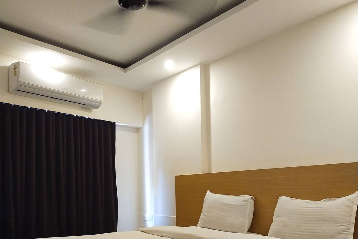 A neatly made bed in a hotel room with a wooden headboard, an air conditioning unit on the wall, and a ceiling fan.
