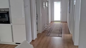 A spacious kitchen with white cabinetry and a built-in oven. A light-colored sofa is in the foreground, and a hallway leads to a door at the end. - Ferienwohnung Direkt in Wehrheim (Wehrheim)