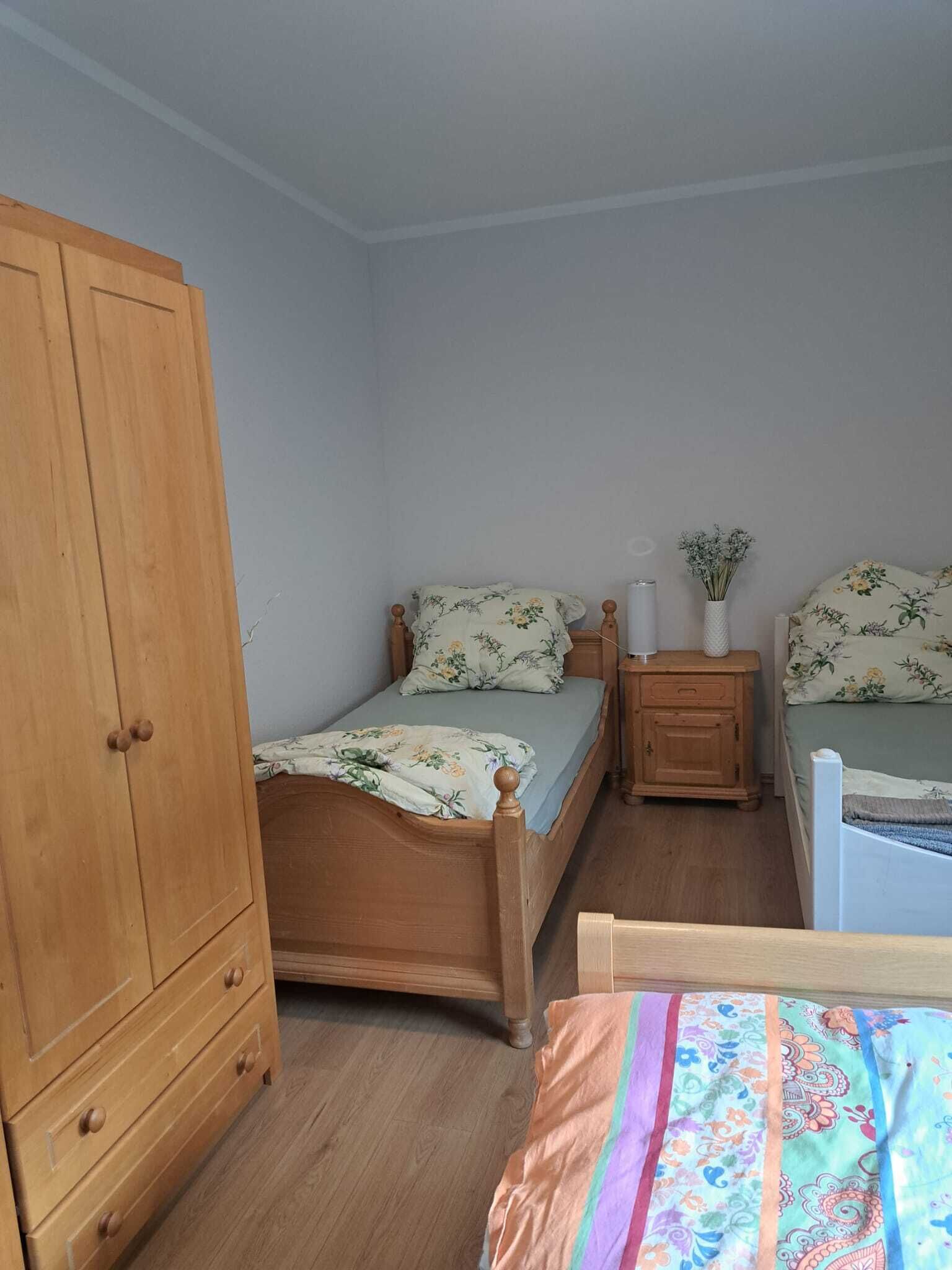 Een slaapkamer met een houten garderobe, een enkel bed met bloemenbeddengoed, een nachtkastje met een vaas bloemen en een klein houten kastje.