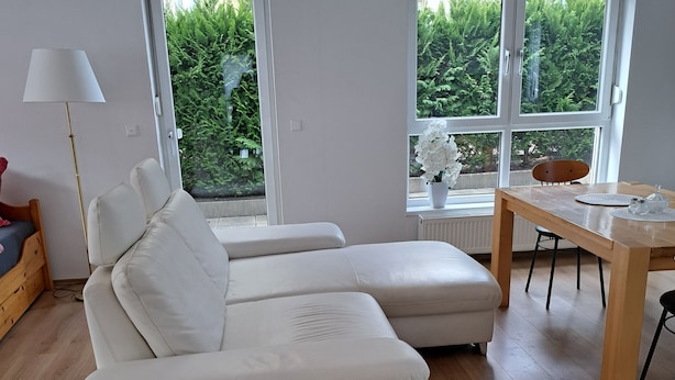 A modern living room with a white sectional sofa, a wooden dining table, and large windows offering a view of greenery. - Ferienwohnung Direkt in Wehrheim (Wehrheim)