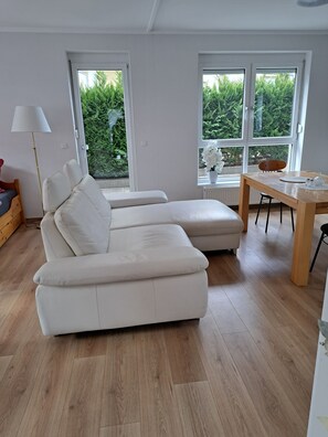 A modern living room with a white sectional sofa, a wooden dining table, and large windows offering a view of greenery. - Ferienwohnung Direkt in Wehrheim (Wehrheim)