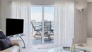 A modern living room with a flat-screen TV, a glass coffee table, a sofa, and a view of a balcony with chairs. - Harbor Central Penthouse (Destin)