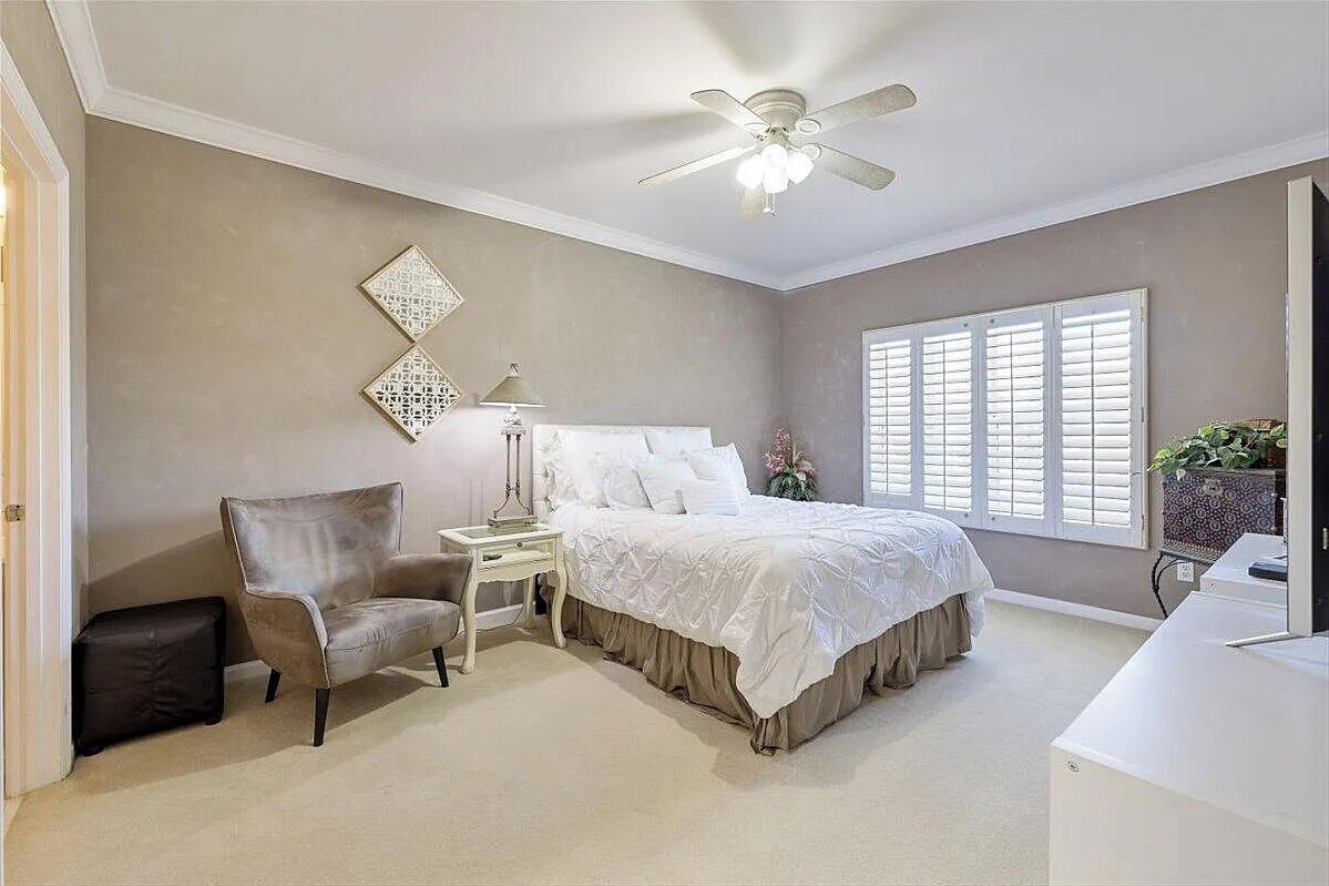 Een slaapkamer met een bed, een stoel, een nachtkastje, een lamp, een plafondventilator en een raam met luiken.