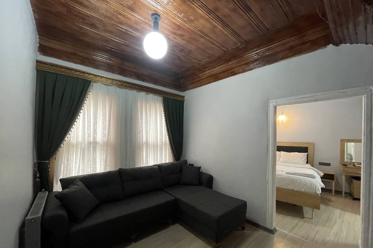 Ein modernes Schlafzimmer mit Holzbalkendecke, einem dunkelgrauen Sofa, einem Bett mit weißer Bettwäsche und einem Nachttisch mit Lampe.