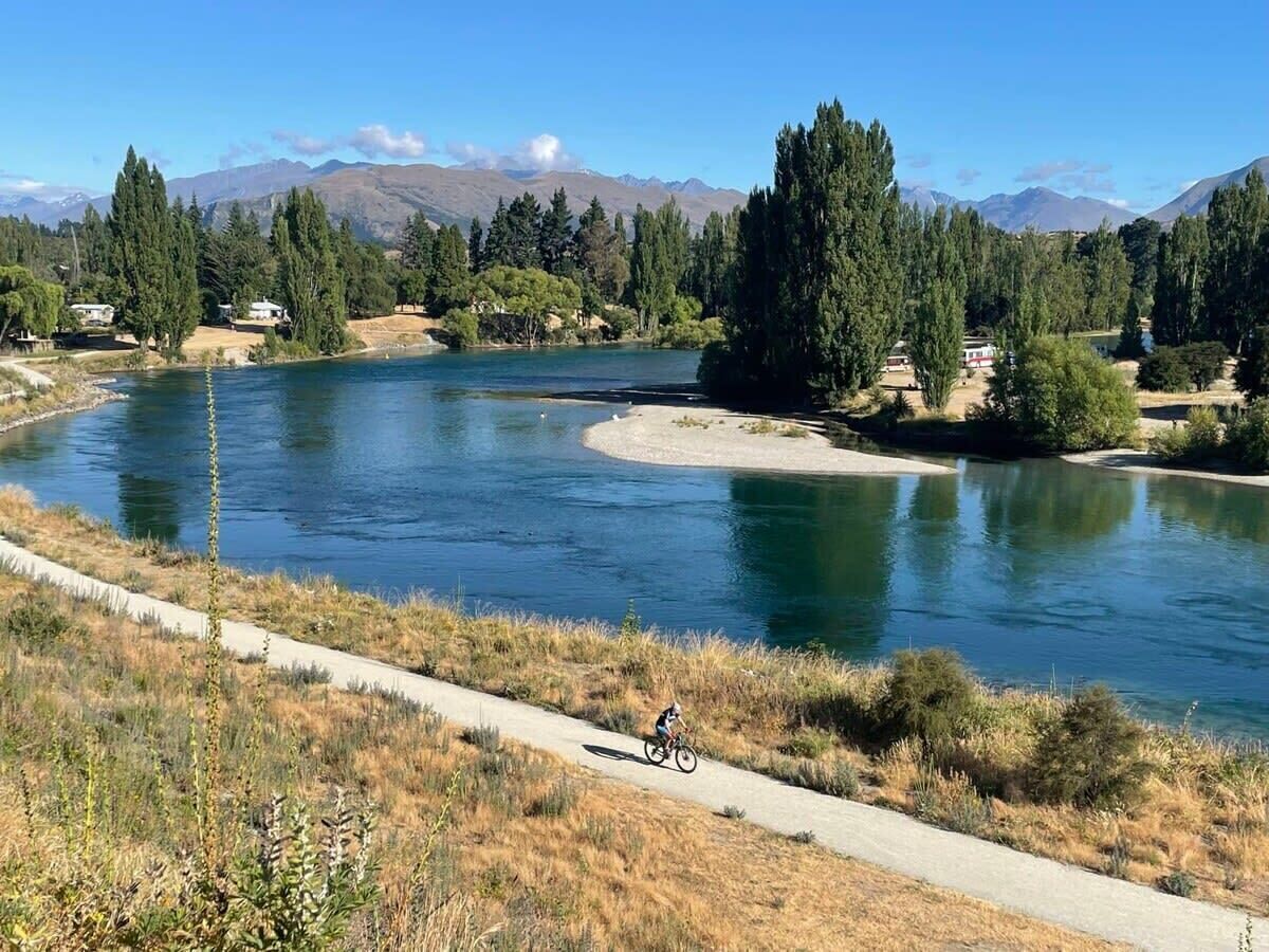 A river with a cyclist on a paved path, surrounded by trees and mountains in the background.