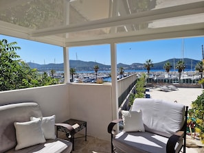A balcony with a white sofa, a small table, and a chair, overlooking a marina with boats and palm trees.