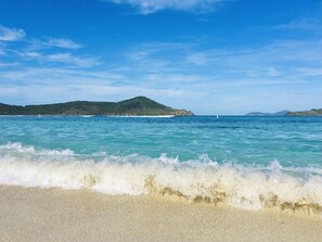 Uma praia com águas turquesa cristalinas, uma faixa de areia e colinas verdes ao longe.