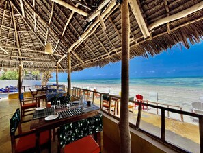 Un restaurante frente al mar con techo de paja, mesas y sillas de madera, con vista a una playa de arena y al océano.