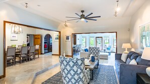 A spacious living room with a gray sofa, patterned armchairs, and a coffee table. There is a dining area with a table and chairs, and a kitchen visible in the background. - Private Luxury Waterfront Villa with Pool (West Bay)