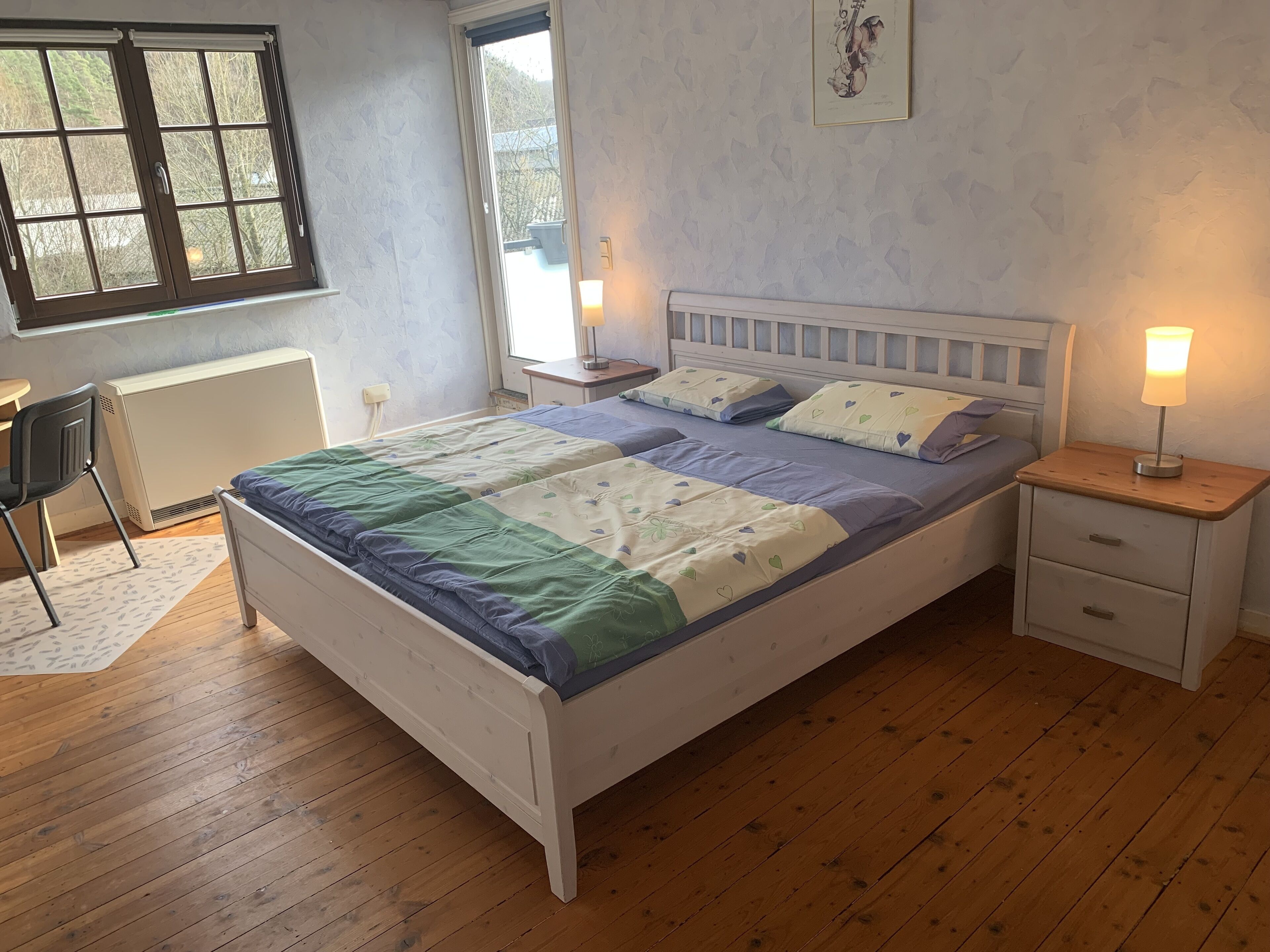 Un dormitorio con cama, mesitas de noche, un radiador, una ventana y una silla.