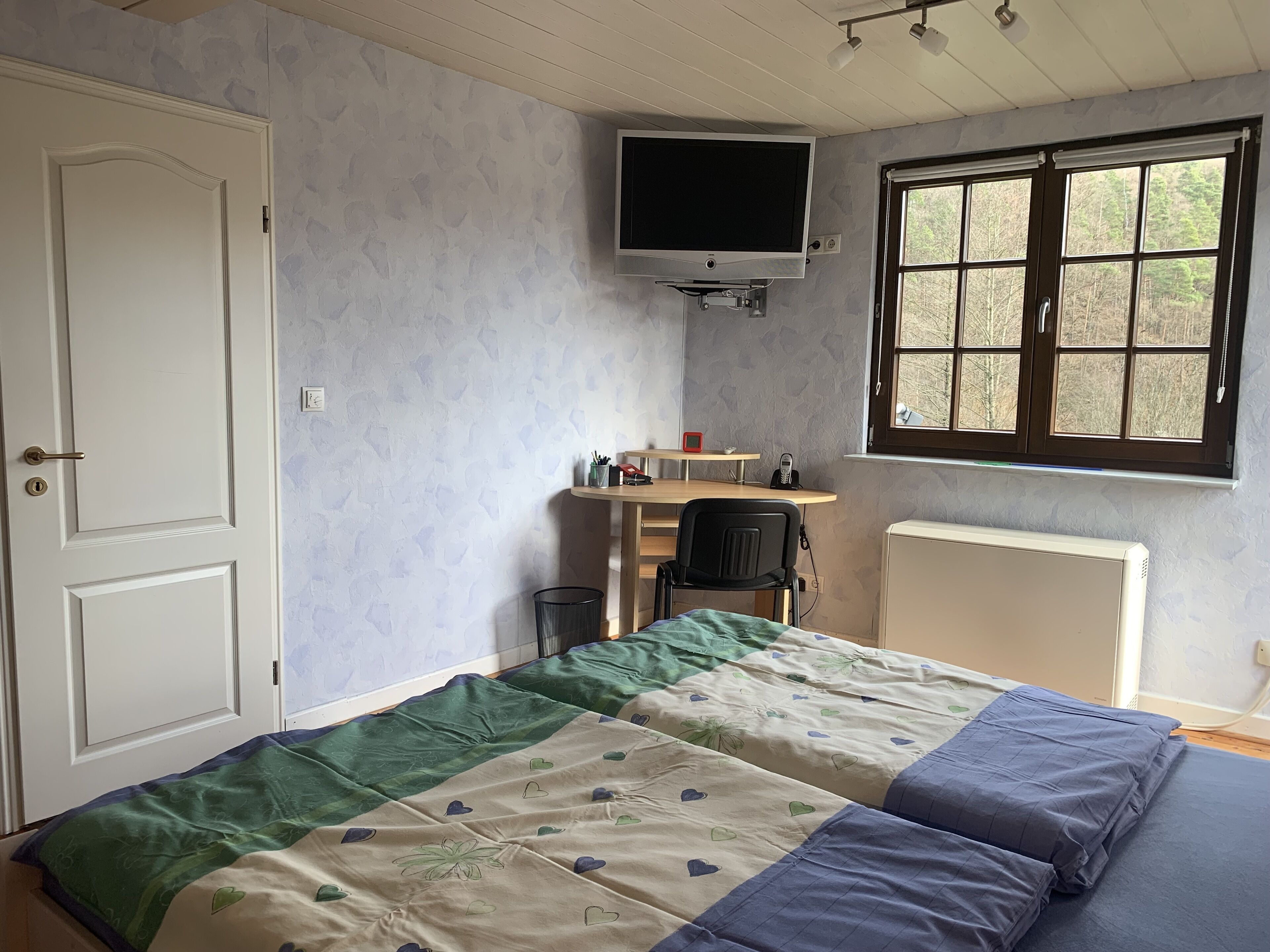 A bedroom with a bed, a desk, a chair, a TV, and a window.