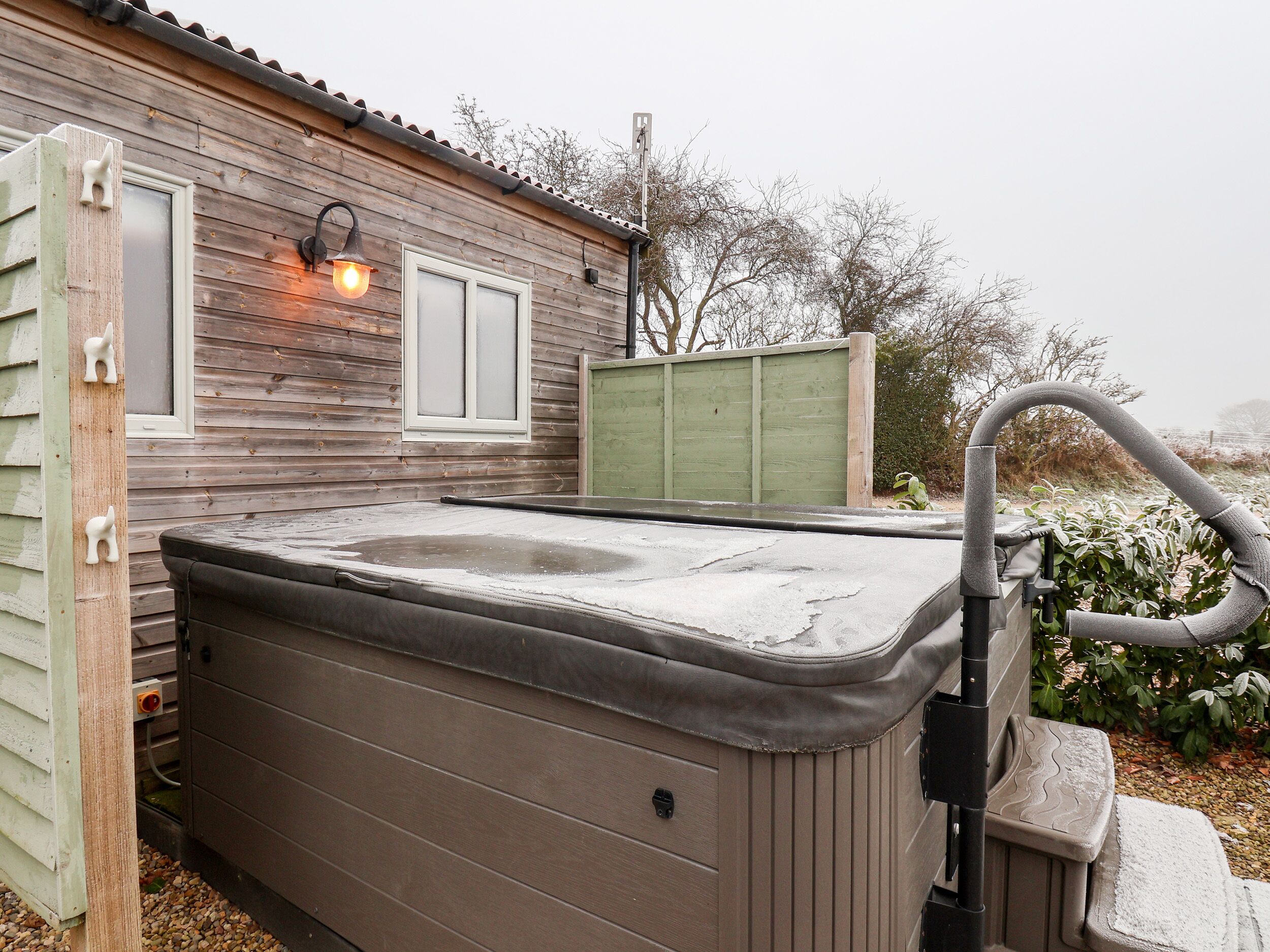 Een hottub voor een houten huis met een raam en een groene hek achtergrond.