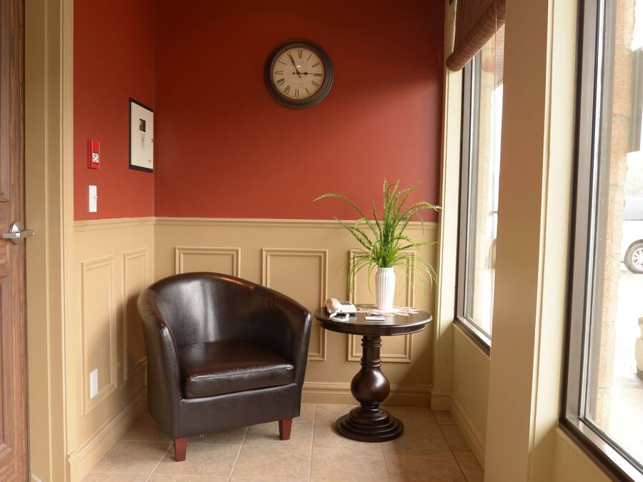 Una habitación con un sillón de cuero oscuro, una pequeña mesa redonda con un jarrón y una planta, un reloj de pared y una ventana con vistas al exterior.
