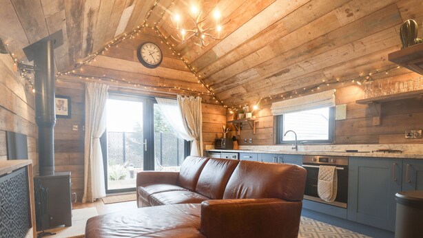 A cozy cabin interior with a leather sofa, wooden walls, a kitchen with blue cabinets, and a large window.